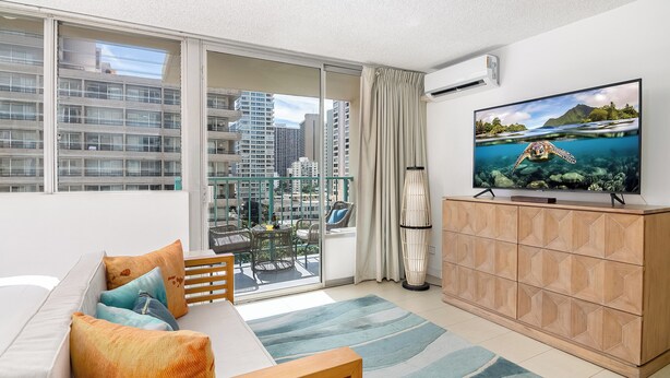 A modern living room with a large TV, a sofa with cushions, a dining table, and a view of the city through large windows. - Studio with King size bed & Short walk to Beach (Honolulu)