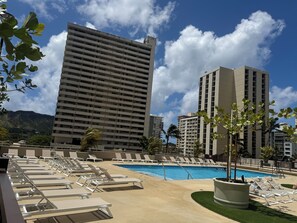 A hotel pool area with sun loungers, a swimming pool, and tall buildings in the background.