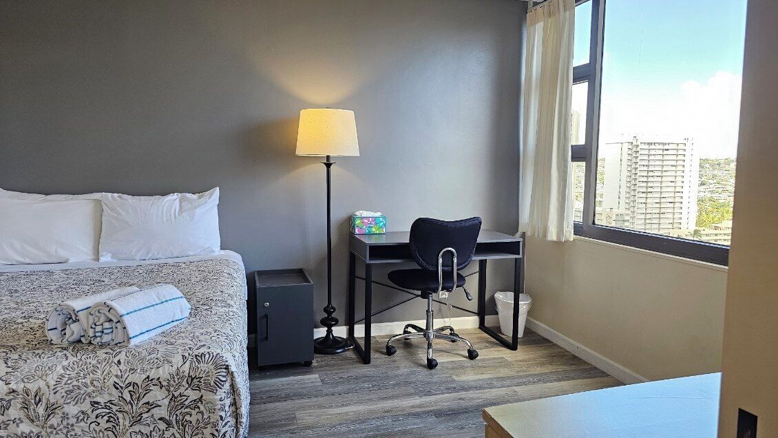 A hotel room with a bed, a desk, a chair, a lamp, and a view of buildings through the window.