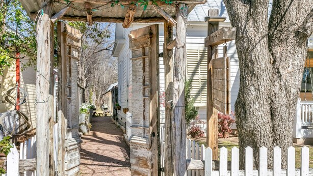 A rustic wooden gate leading to a pathway surrounded by trees and a white picket fence. - Centre St. Cottage | 1/1, King Bed, Mini Kitchen (Fredericksburg)