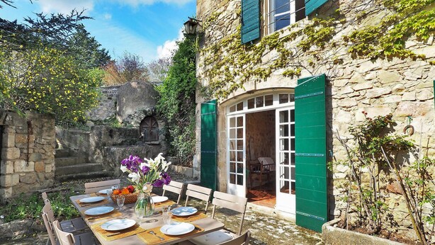 Un espacio para comer al aire libre con pared de piedra, contraventanas verdes y una mesa dispuesta para cuatro.