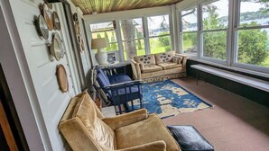 A cozy living room with a wooden ceiling, large windows, a beige sofa, a blue armchair, and a patterned rug. - Lakefront house with land dock (Conneaut Lake)