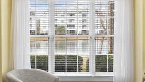 A room with a large window featuring horizontal blinds, a grey armchair, and a view of buildings and trees.