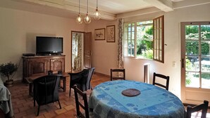 Un comedor con una mesa redonda cubierta con mantel azul, rodeada de sillas de madera. Un televisor está montado en un mueble de madera a la izquierda. La habitación tiene ventanas grandes con cortinas, que permiten luz natural y vistas de la vegetación exterior.