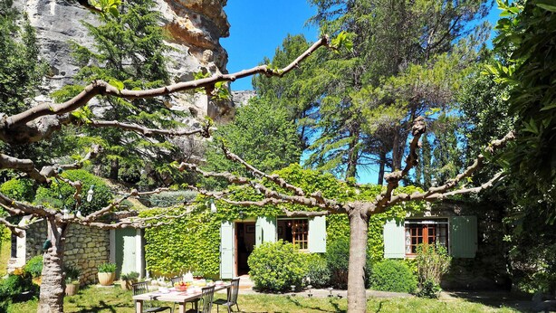 Un edificio rústico de piedra con un jardín, un comedor con mesa y sillas, y un árbol grande con una rama extendida.