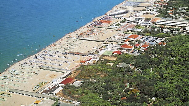 An aerial view of a coastal area with a beach lined with rows of beach umbrellas and sun loungers. There are buildings adjacent to the beach, and a forested area to the right. - Le Dune by Interhome (Tirrenia)