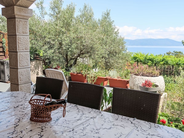 A terrace with wicker chairs and a table, surrounded by potted plants and a view of the sea.