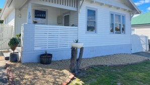 A white single-story house with a covered porch, a small front yard with a tree and potted plants, and a mailbox. - Comfy home within walking distance to beach, restaurants and Blunderstone area. (Bellerive)