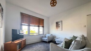 A living room with a flat-screen TV, a grey sofa, a white armchair, and a wooden cabinet. - Comfy home within walking distance to beach, restaurants and Blunderstone area. (Bellerive)