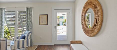 A room with a wooden floor, a striped sofa, a woven bench, a framed picture on the wall, and a door leading outside.