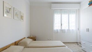 Ein Schlafzimmer mit zwei Betten, einem Fenster mit durchsichtigen Vorhängen, einem Nachttisch mit Lampe und einem Kleiderschrank.