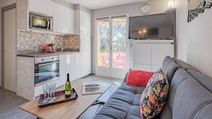 Una cocina moderna con microondas, horno y salpicadero de azulejos. Hay una zona de comedor con una mesa y un sofá. Un televisor está montado en la pared y una ventana ofrece vistas al exterior.