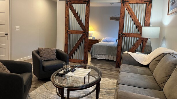 A modern living room with a glass coffee table, two sofas, a bed with striped curtains, and a sliding barn door.