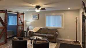 A living room with a brown leather sofa, a glass coffee table, a ceiling fan, and a large window with blinds.
