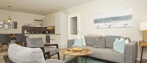 Ein modernes Wohnzimmer mit einem grauen Sofa, zwei Sesseln, einem Couchtisch und einer Küchenzeile im Hintergrund.
