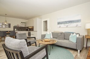 Una sala de estar moderna con un sofá gris, dos sillones, una mesita de centro y una cocina al fondo.
