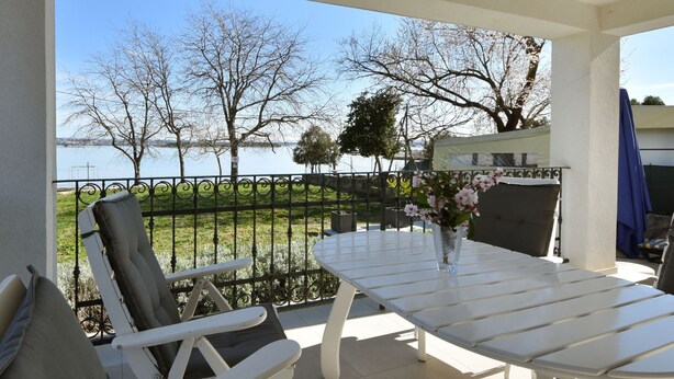 A balcony with a white table and chairs, a vase with flowers, and a view of a lake and trees. - Sea side by Interhome (Pašman/Kraj)