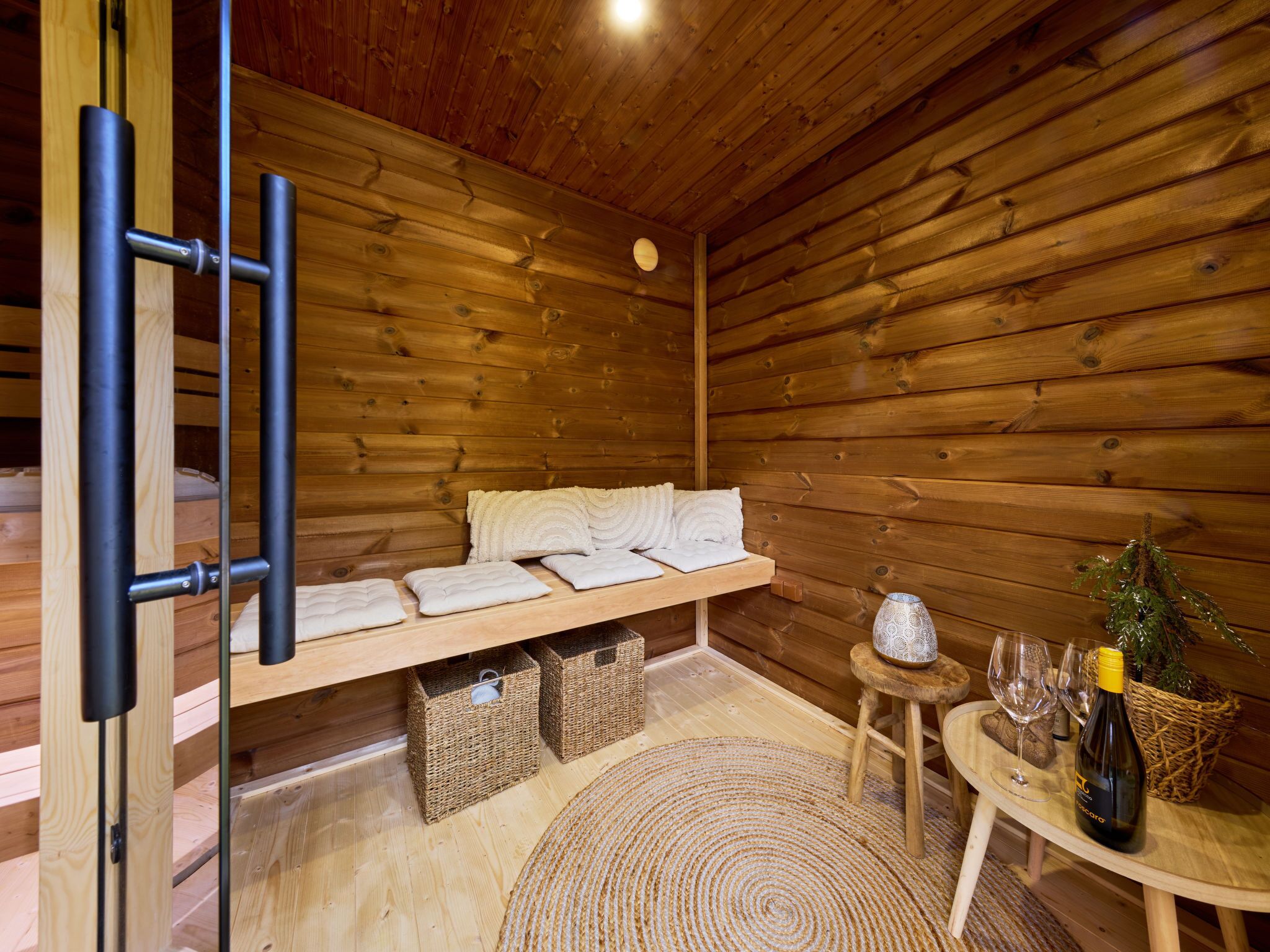 L’intérieur d’un chalet en bois, avec un banc en bois, des serviettes, des paniers, un tapis tissé, une petite table avec une bouteille de vin et des verres.