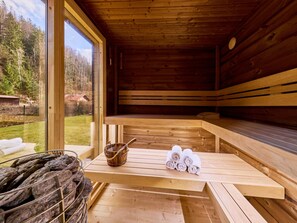 Uma sauna de madeira com bancos, uma mesa com toalhas e um balde, e vista para uma área arborizada.