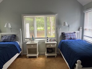 A bedroom with two beds, each with blue bedding and plaid pillows. There are white nightstands with lamps and a window with blinds.