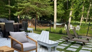 Una terraza al aire libre con mobiliario de madera, incluyendo sillas y una mesa, rodeada por un área cercada con árboles.