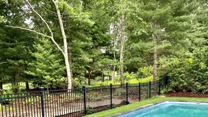 Una piscina con borde de piedra y borde de césped, rodeada por una valla negra y árboles.