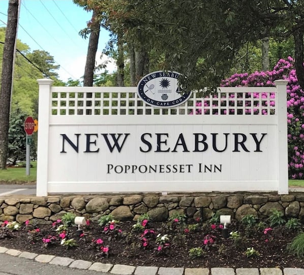 Señal para el New Seabury Popponeset Inn, con fondo blanco y letras negras, rodeado de un muro de piedra y plantas con flores.