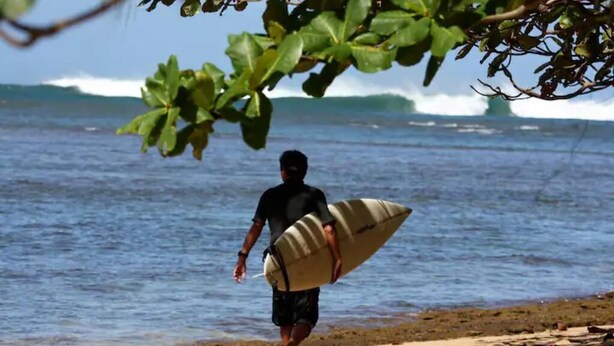 Een persoon met een surfplank die over het strand loopt, met golven op de achtergrond.