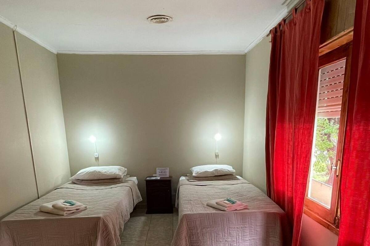 A hotel room with two single beds, a small table, and a window with red curtains.