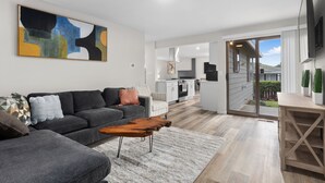A modern living room with a grey sofa, a wooden coffee table, and a large abstract painting. There is a kitchen area visible through an open door, and a dining table with chairs. - Cozy 3-bedroom house in peaceful Renton with WiFi, AC (Renton)