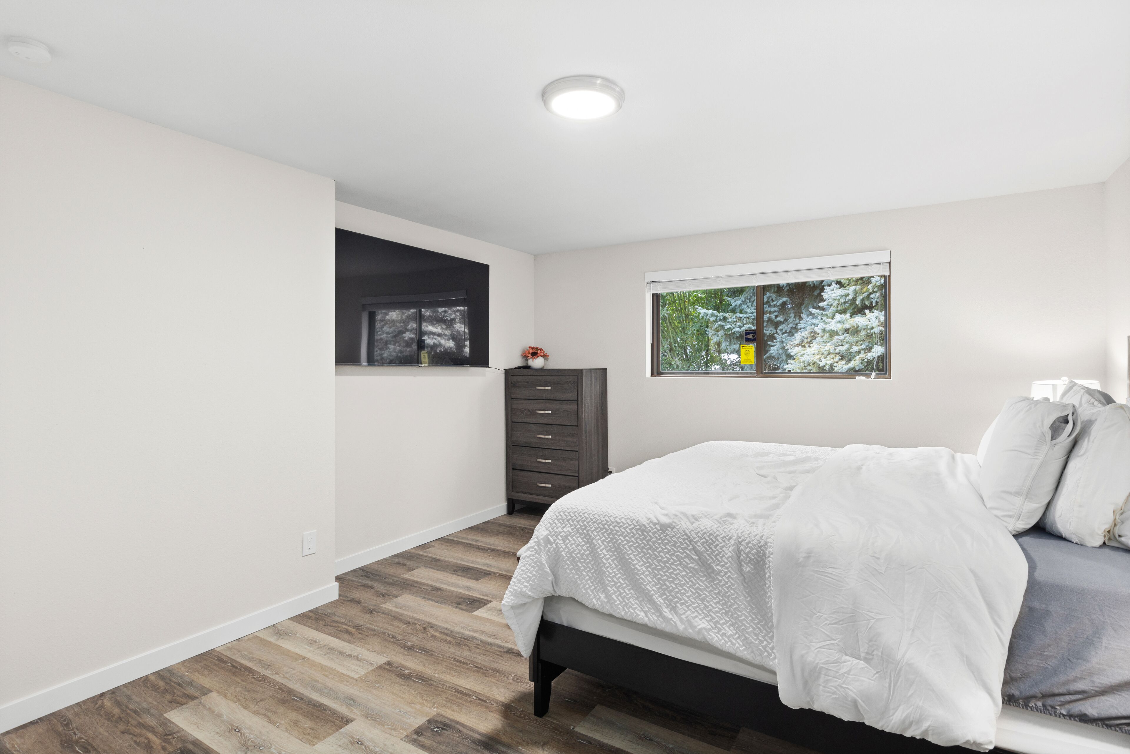 Une chambre à coucher avec un lit, une commode, une fenêtre donnant sur des arbres et un luminaire au plafond.
