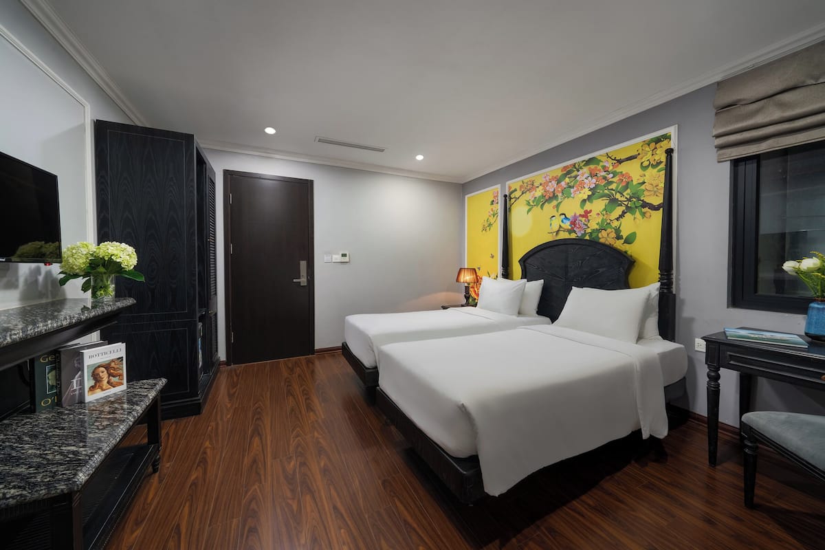 Une chambre d’hôtel avec deux lits, un téléviseur, un plancher en bois, un tableau au mur et un bureau avec une lampe.