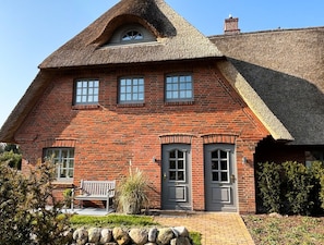 A traditional brick house with a steeply pitched thatched roof, a front porch with a bench, and a stone wall in the foreground. - Südfall - Ein Kuschelnest für Zwei Erwartet Sie in Tinnum (Tinnum)
