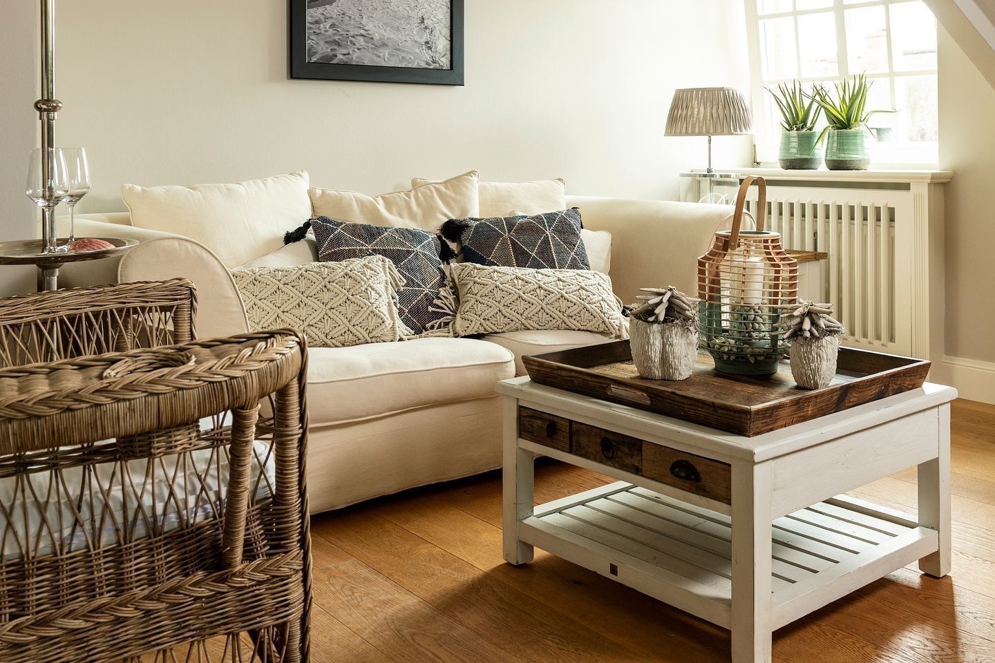 A cozy living room with a white sofa, patterned cushions, a wicker chair, a wooden coffee table with decorative items, and a window with plants.