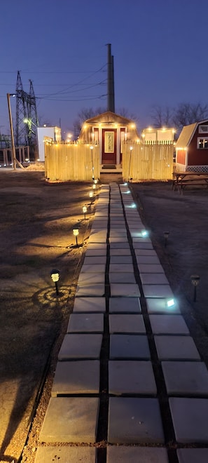 A pathway leading to a small building with a red door, surrounded by a wooden fence and illuminated by string lights. - Cozy cabin on a working horse rescue with WiFi, HVAC, plumbed outhouse, shower (Coweta)