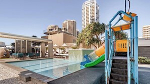 Uma piscina com escorregador, espreguiçadeiras e um horizonte urbano ao fundo.