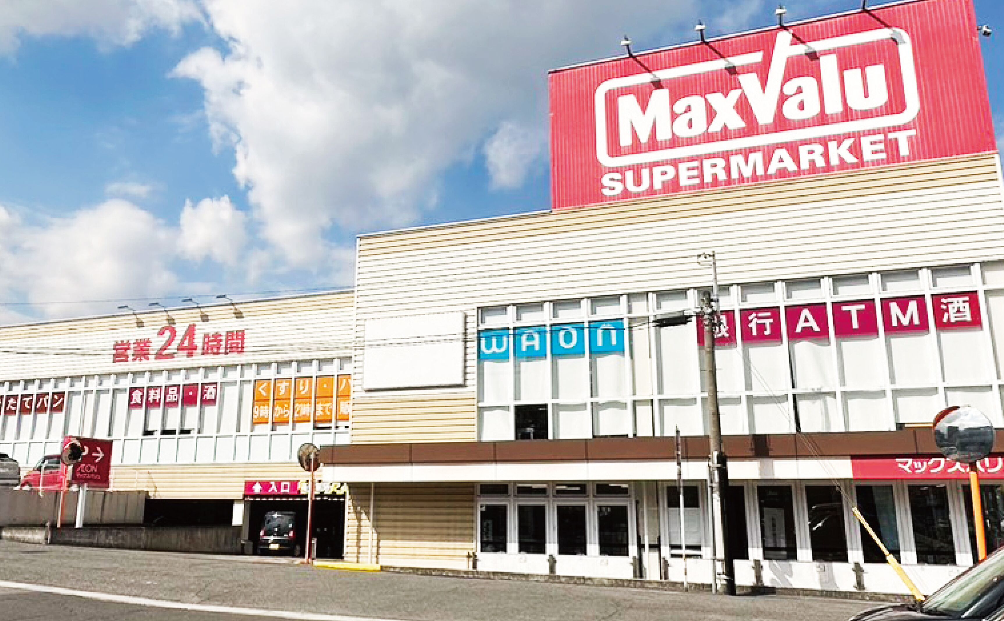 Un supermarché MaxValu avec une enseigne indiquant qu’il est ouvert 24 heures sur 24, un distributeur automatique de billets et une entrée bien visible.