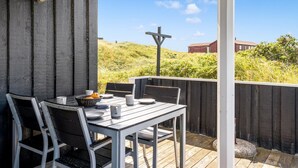 Un espace extérieur abrité, aménagé pour prendre les repas, avec une table et des chaises, un panier de fruits, et une vue sur une colline et des bâtiments en arrière-plan.