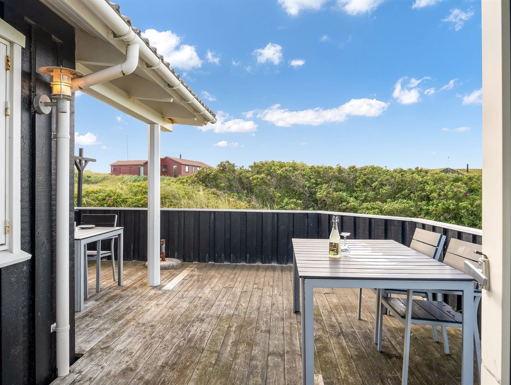 A wooden deck with a table and chairs, a pergola, and a view of a red building and greenery in the background.