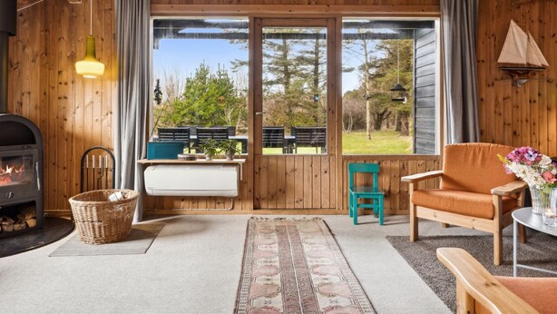 A cozy cabin interior with a fireplace, a dining table, a sofa, and a large window offering a view of the outdoors.