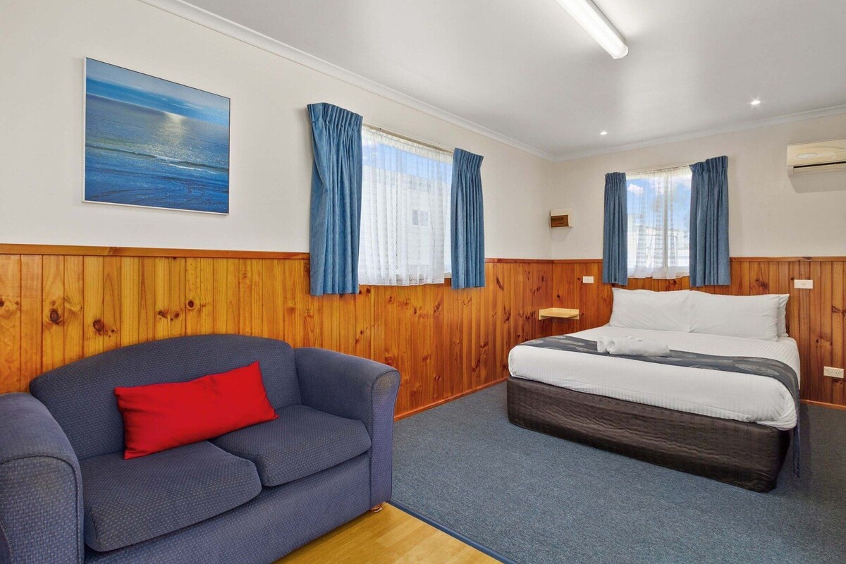 Una habitación con cama, un sofá azul con un cojín rojo, paneles de madera, un cuadro de una escena marina y una ventana con cortinas azules.