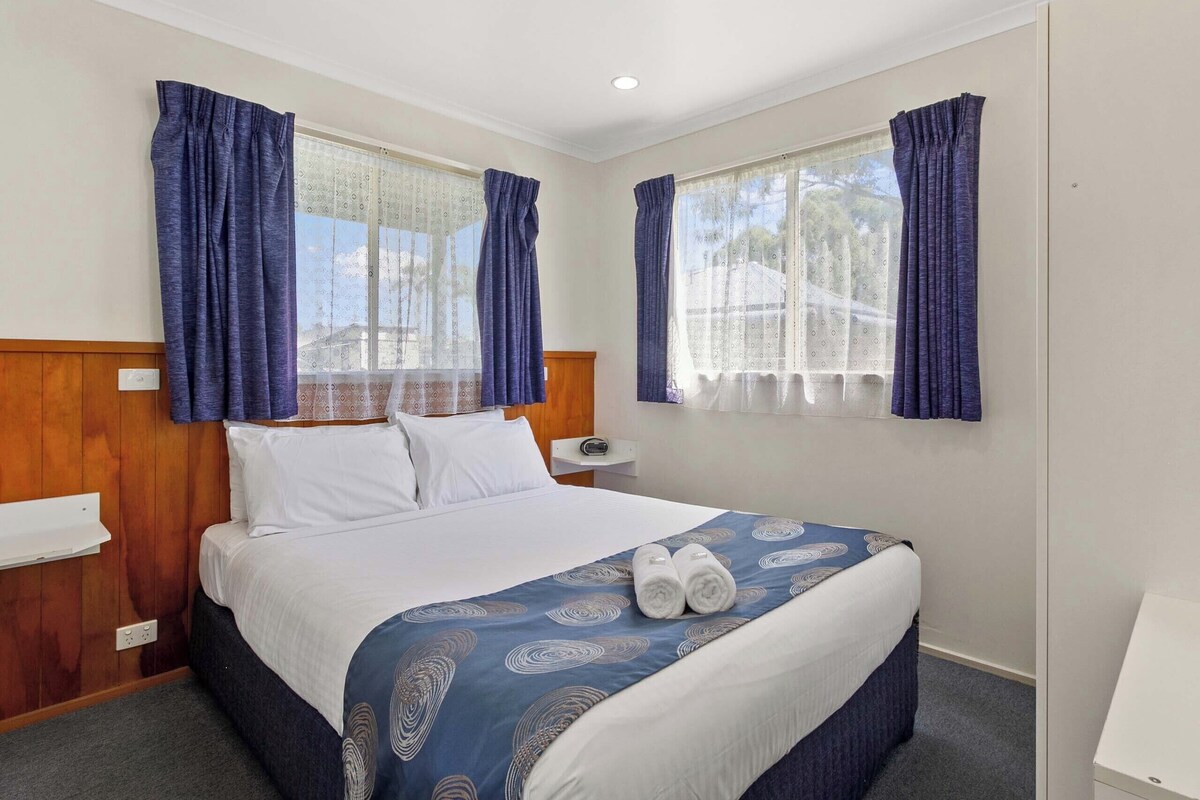 Habitación de hotel con cama, dos ventanas con cortinas, una mesita de noche y una lámpara fijada a la pared.