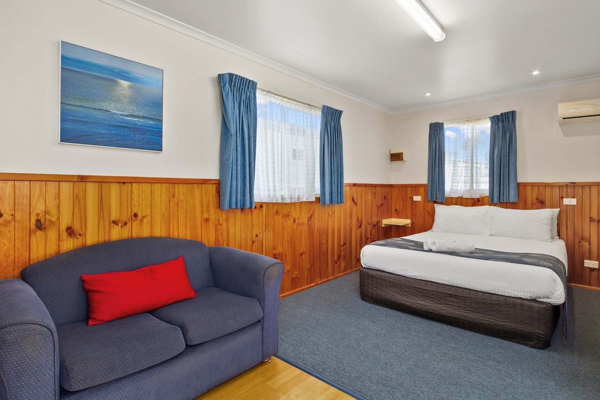 Een kamer met een bed, een blauwe bank met een rood kussen, houten lambrisering, een schilderij van een zeegezicht en een raam met blauwe gordijnen.