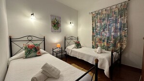 Two single beds with wooden headboards, white linens, and floral pillows. There are bedside tables with lamps and a colorful tapestry on the wall. - Quinta Jardim das Palmeiras, T2 nº3, Algoz (Silves)