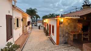 A narrow cobblestone street with buildings on either side, featuring outdoor seating areas and a brick fireplace. - Quinta Jardim das Palmeiras, T2 nº6, Algoz (Silves)