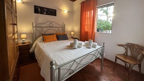 Un dormitorio con estructura de cama metálica, techo de madera, una ventana con cortinas naranjas y una silla.