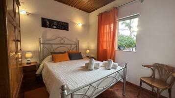 Un dormitorio con estructura de cama metálica, techo de madera, una ventana con cortinas naranjas y una silla.