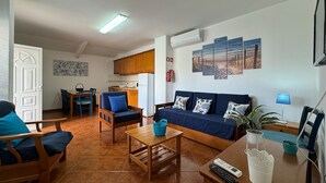 Un salon avec un canapé, des fauteuils, une table basse et un téléviseur. Au fond, on aperçoit une cuisine avec des placards et un réfrigérateur.
