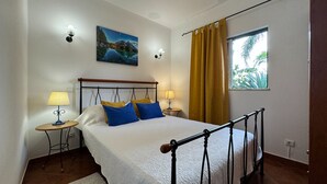 Une chambre à coucher avec un lit, deux tables de chevet, une commode, un tableau, une fenêtre avec des rideaux et une vue sur la verdure à l’extérieur.