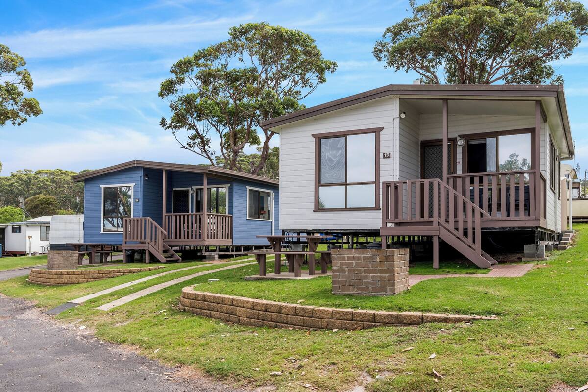 A mobile home with a porch and a small deck, surrounded by a grassy area and trees.